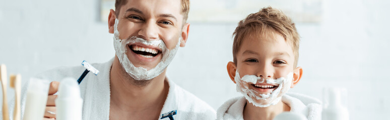 horizontal image of happy father and son with shaving foam on faces holding shaving razors