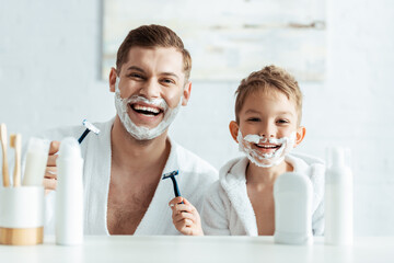 selective focus of cheerful father and son with shaving foam on faces holding shaving razors