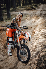 Fototapeta na wymiar Charming young female racer with semi naked torso sitting on her bike in the woods, smiling and lokking on the camera