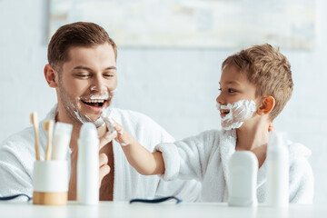 selective focus of smiling boy applying shaving foam on face of cheerful father