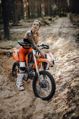 Tattooed female racer wearing motocross outfit with semi naked torso sitting on her bike in the forest