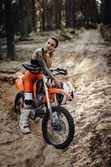 Obraz na płótnie Canvas Beautiful female racer wearing motocross outfit with semi naked torso sitting on her bike in the forest
