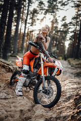 Fototapeta na wymiar Cheeky tattooed racer girl wearing motocross outfit with semi naked torso sitting on her bike at the bright sunset in the forest