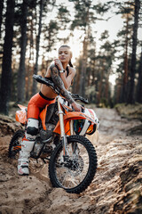 Fototapeta na wymiar Cheeky tattooed racer girl wearing motocross outfit with semi naked torso sitting on her bike at the bright sunset in the forest