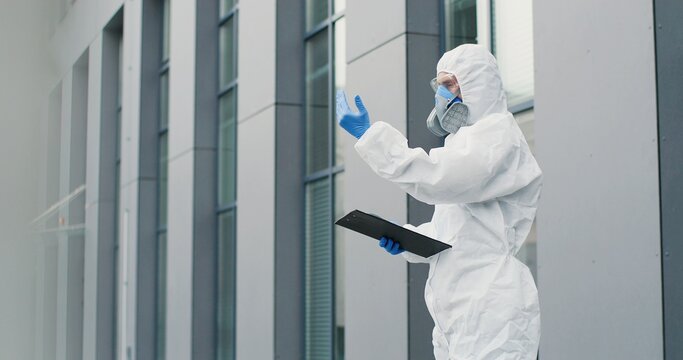Female office worker in mask going to work and man in white suit and respirator measuring her temperature, letting go. Allowing pass to business center for visitor. Covid-19 pandemic. Permit to enter.
