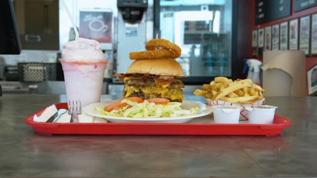 Woman employee at a burger joint pushes a full tray with the largest burger and fries and shake across the counter for the customer to pick up.
