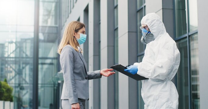Female office worker in mask going to work and man in white suit and respirator measuring her temperature, letting go. Allowing pass to business center for visitor. Covid-19 pandemic. Permit to enter.