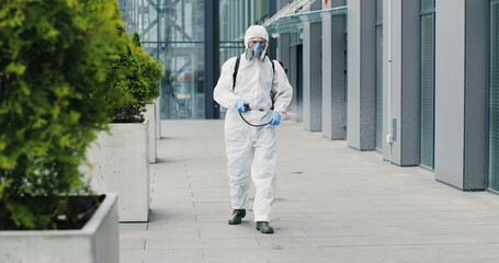 Caucasian man in special antibacterial white suit and respirator walking the street and disinfecting sidewalk with pulverizator. Spraying disinfectant on ground outdoors. Coronavirus pandemic concept.