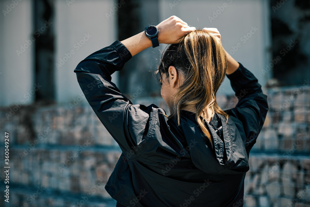 Wall mural Rear view of attractive fit sportswoman in hooded shirt tying hair and preparing for running.