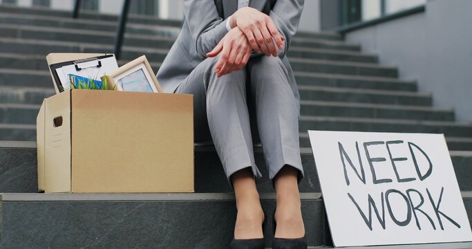 Portrait of Caucasian young workless woman siting on stairs at street with carton board Need Work and box with stuff. Female fired office worker in depression. Unemployment concept.