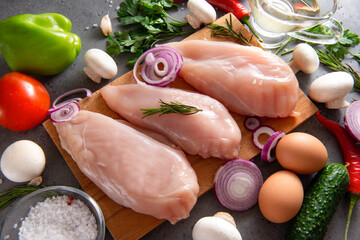 pieces of raw chicken fillet on a cutting Board, meat, vegetables, herbs, tomato, onion, parsley, oil, pepper, eggs, cucumber, mushrooms, salt on a gray background