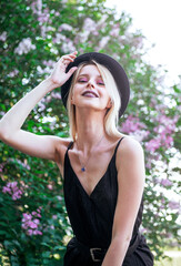 stylish blonde with a black hat on a background of blooming lilac, a young white woman smiling