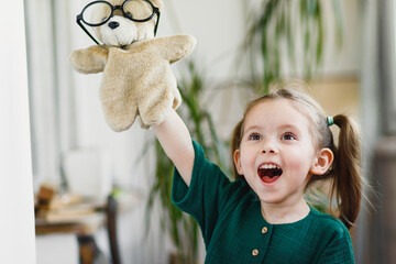 Sweet little girl is playing with a teddy bear in round shaped glasses and smiling while stay at...