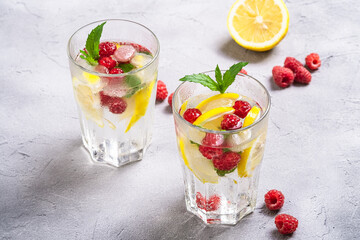 Fresh cold ice water with lemon, raspberry fruits and mint leaf in two faceted glass on stone concrete background, summer diet beverage, angle view