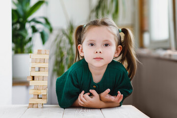 Cute and serious caucasian little girl with ponytails sitting at a table near by a wooden tower and looking at a camera