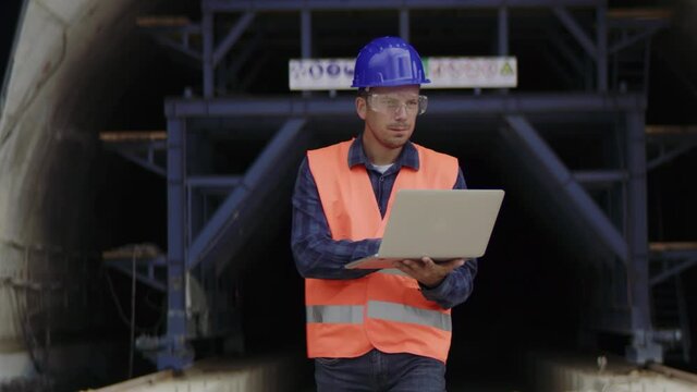 Construction engineer, architect or a supervisor wearing a protective equipment using a computer while exiting an underconstruction tunnel.