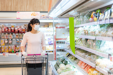 Asian woman with hygienic mask and rubber glove with shopping cart in grocery and looking for fresh vegetable pack to buy during covid-19 outbreak for preparation for a pandemic quarantine