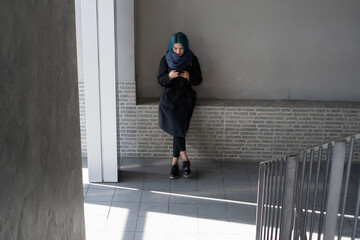 A girl in a black coat and blue scarf is typing on the phone while standing against a white wall.
