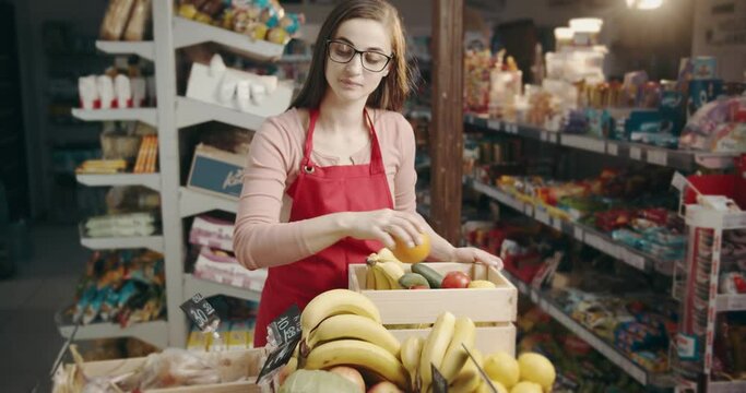 Beautiful female worker in glasses and apron restocking fresh fruits and vegetables into boxes at store. Competent assistant with dark hair controlling fresh food at supermarket