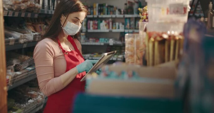 Saleswoman in protective mask and gloves using digital tablet while doing revision of food at supermarket. Responsible girl keeping quarantine regulations while working at grocery store.