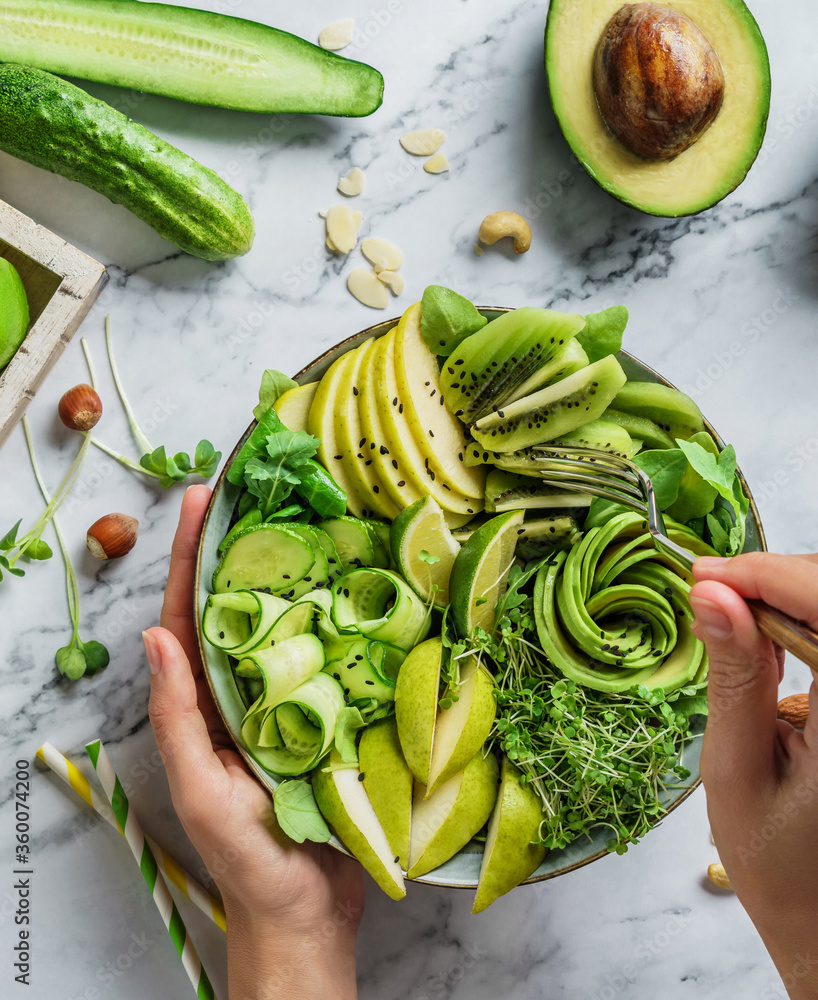 Wall mural hands holding healthy fresh summer salad with avocado, kiwi, apple, cucumber, pear, greens and sesam