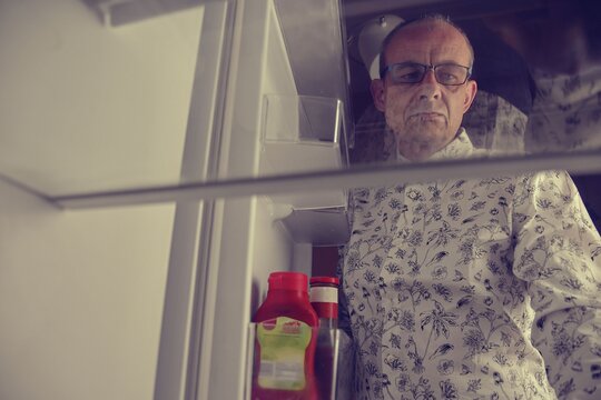 Hungry Man Looking For A Late Night Meal In An Empty Fridge. Tired Old Man Looking For A Snack In The Fridge At Night, View From The Fridge