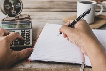 close up of a businessman who writes or draw notes in a blank notebook Pressing calculator On a wooden office desk ready to drink coffee in the morning.