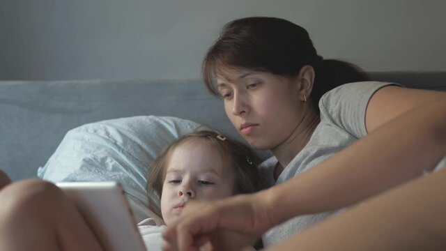 Mom and little daughter are lying in bed with wake cartoon on a tablet. mother and baby make video call to father. Technology concept, new generation, family, connection, parenthood