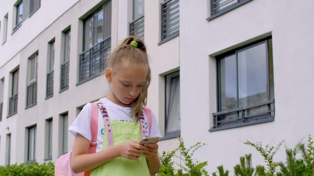 Focused teenage girl walking on street and using smartphone. Adorable concentrated teen girl with pink backpack walking on urban city street and using mobil phone