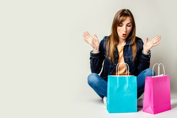 Young woman with a surprised face in a denim jacket holds shopping bags while sitting on the floor on a light background. Banner. Concept shopping, sale, discounts