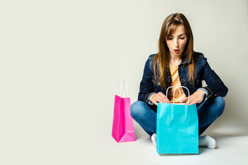 Young woman with a surprised face in a denim jacket holds shopping bags while sitting on the floor on a light background. Banner. Concept shopping, sale, discounts