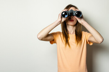 Young friendly girl in a T-shirt looks through binoculars on a light background