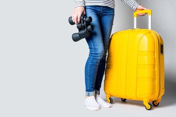 Woman holds a plastic yellow suitcase and binoculars on a light background. Travel concept, flight expectation, vacation. Only legs are visible. Banner