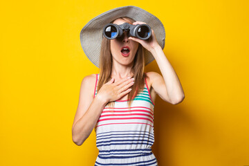 Young woman in shock in surprise with a hat and a striped dress looks in surprise with binoculars on a yellow background