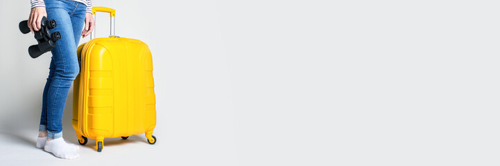 Fototapeta premium Woman holds a plastic yellow suitcase and binoculars on a light background. Travel concept, flight expectation, vacation. Only legs are visible. Banner