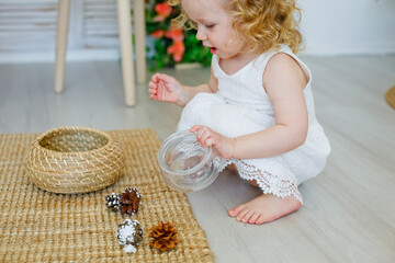 A little girl smiles and plays with a pine cone. A cute young blonde girl is studying in a classroom in the style of Montessori or home concept. Explores natural materials.