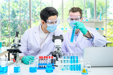 Couple male scientist wearing protection suit holding beaker or tube with sample liquid inside and working with many lab equipment for research at laboratory.