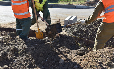 Workers throw earth with construction shovels, dig a hole for road curbs.