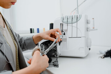 Young woman tailor is cutting with scissors a piece of black material in her white mini workshop.