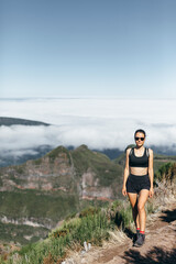 Naklejka premium Young athletic woman in black sports top and shorts walks along a trail in mountains. Bottom panorama of clouds and peaks. 