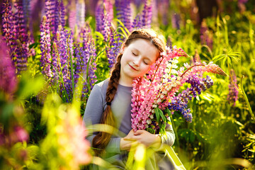 Close-up portrait of a cute little girl with a bunch of lupins in the sunlight. A girl with closed eyes holds a bouquet of purple flowers in a field of lupines. Child with a bouquet