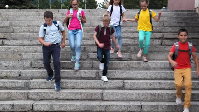 group of happy junior schoolchildren with backpacks run down large stone stairs together on sunny summer day slow motion