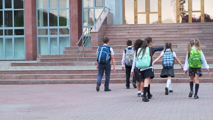junior students in uniform with schoolbags walk to school building with stone steps on warm day backside view slow motion