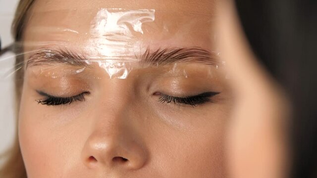Create permanent eyebrow makeup. Microblading eyebrows work flow in a beauty salon. Cosmetician putting on film on eyebrows. Permanent makeup for eyebrows. Eyebrow lamination and styling
