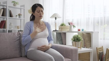 Pregnant woman suffer from nausea feeling while resting in comfort couch in home living room. illness relax asian chinese future motherhood having morning sickness and cover mouth with hands.