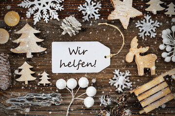 One White Label With German Text Wir Helfen Means We Help. Frame Of Christmas Decoration Like Tree, Sled, Star And Fir Cone. Wooden Background With Snowflakes
