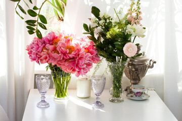 Bouquet of peonies and roses, black and white candles, wine glasses and old tea pot on white table near the window.
