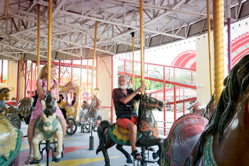 Fototapeta na wymiar Elderly couple spending time together at theme park on weekend, senior people hanging out and having fun at amusement park. Grandpa and grandma enjoying at carousel spinning