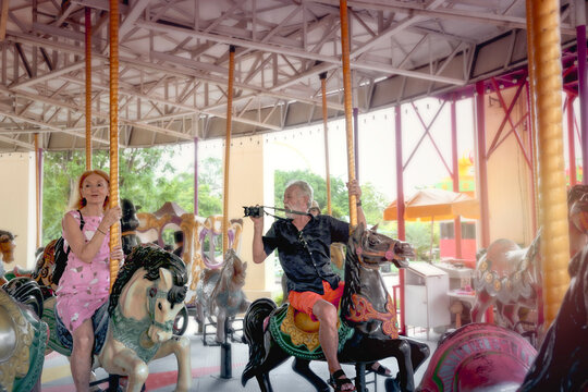 Elderly Couple Spending Time Together At Theme Park On Weekend, Senior People Hanging Out And Having Fun At Amusement Park. Grandpa Taking Photo Of Grandma Enjoying At Carousel Spinning