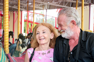 Obraz premium Elderly couple spending time together at theme park on weekend, senior people hanging out and having fun at amusement park. Grandpa and grandma enjoying at carousel spinning